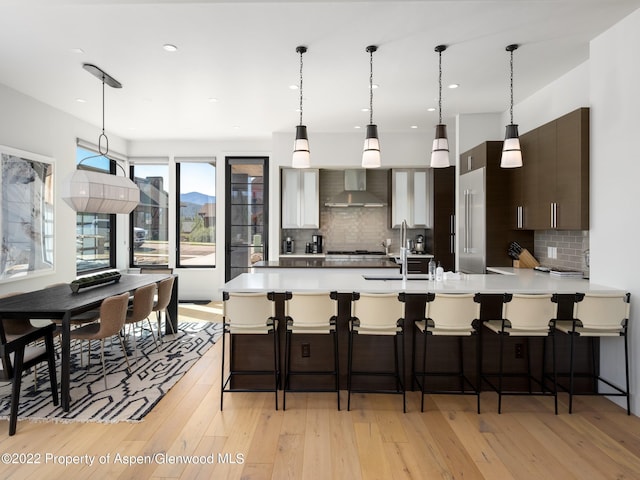 kitchen with light hardwood / wood-style floors, decorative backsplash, dark brown cabinets, wall chimney range hood, and a breakfast bar