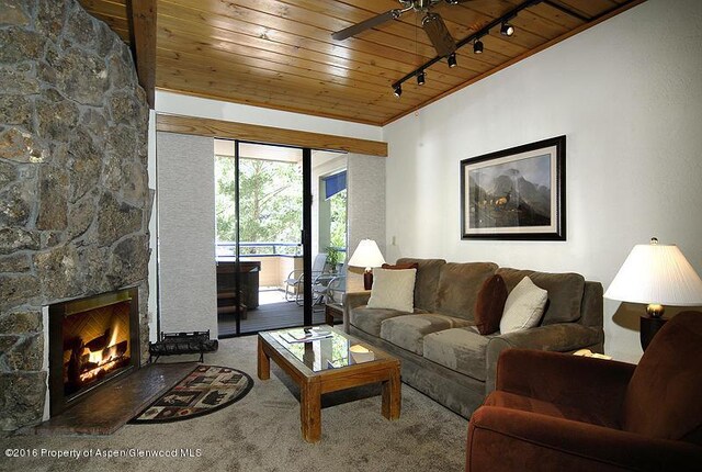 carpeted living room with a stone fireplace, ceiling fan, wood ceiling, and track lighting