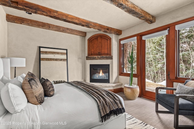 bedroom with a glass covered fireplace, access to outside, light colored carpet, and beam ceiling