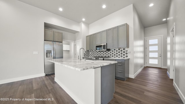 kitchen with decorative backsplash, dark wood-type flooring, a kitchen island with sink, gray cabinets, and stainless steel appliances