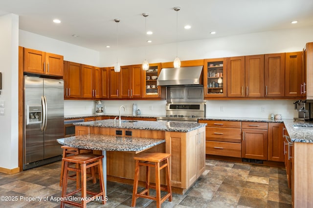 kitchen featuring stone counters, exhaust hood, appliances with stainless steel finishes, an island with sink, and glass insert cabinets