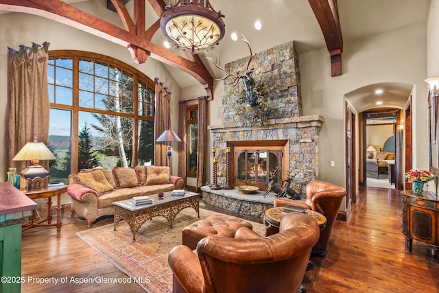 living area with wood finished floors, beam ceiling, a stone fireplace, high vaulted ceiling, and arched walkways