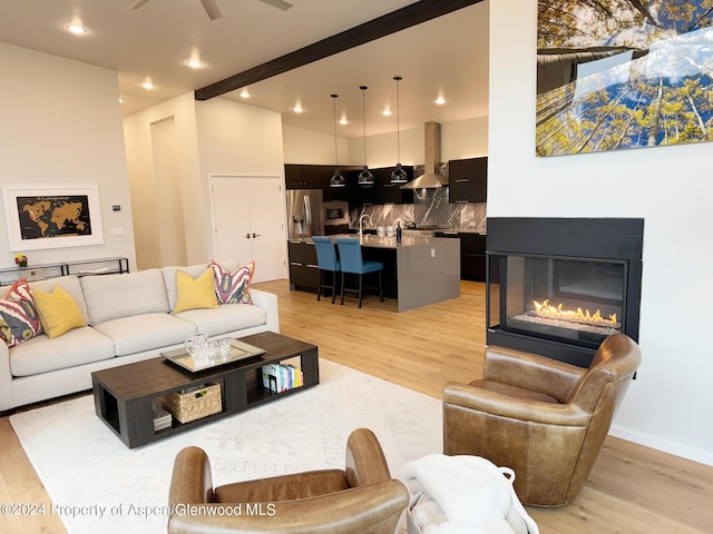 living room with a multi sided fireplace, sink, light hardwood / wood-style flooring, ceiling fan, and beamed ceiling