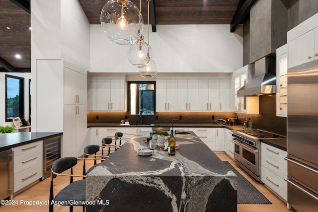 kitchen featuring decorative backsplash, high end appliances, wall chimney range hood, white cabinets, and hanging light fixtures