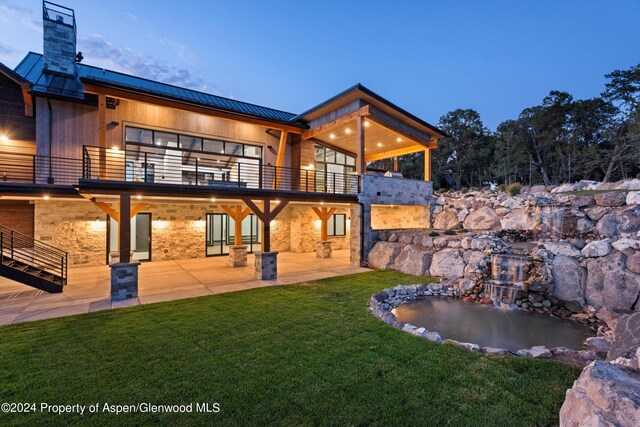 back house at dusk featuring a patio, a balcony, and a lawn