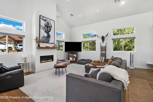 living room featuring high vaulted ceiling, wood finished floors, a glass covered fireplace, baseboards, and recessed lighting