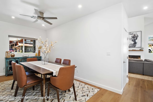 dining space with light wood-type flooring, ceiling fan, a lit fireplace, baseboards, and recessed lighting