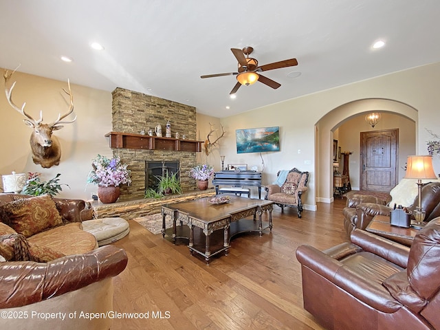 living room featuring arched walkways, wood finished floors, a ceiling fan, a stone fireplace, and recessed lighting