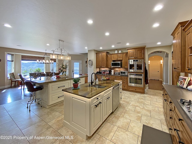 kitchen featuring a sink, a center island with sink, appliances with stainless steel finishes, a kitchen bar, and brown cabinetry