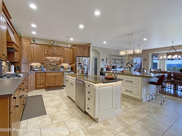 kitchen with an island with sink, appliances with stainless steel finishes, dark countertops, decorative backsplash, and a chandelier