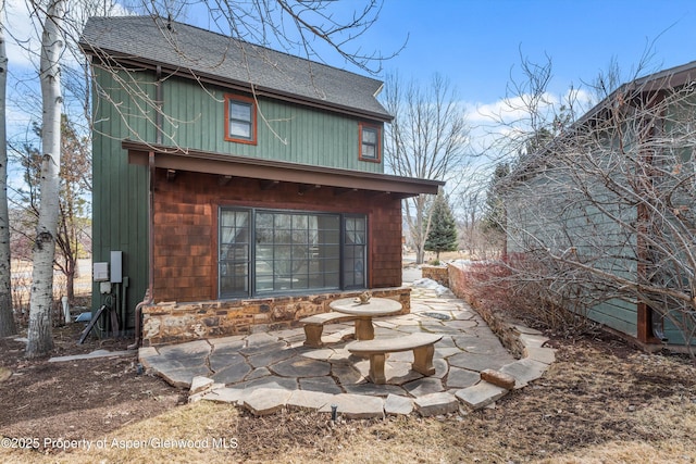 rear view of house with stone siding, a patio, and roof with shingles