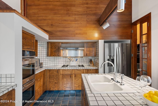 kitchen featuring wall chimney range hood, sink, decorative backsplash, tile counters, and stainless steel appliances