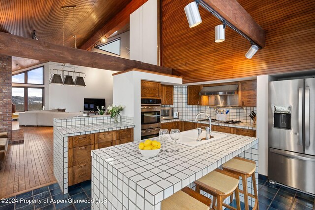 kitchen with decorative backsplash, wall chimney exhaust hood, stainless steel appliances, a kitchen island with sink, and tile countertops