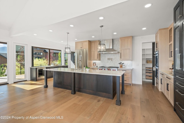 kitchen with pendant lighting, backsplash, a kitchen island with sink, wall chimney range hood, and appliances with stainless steel finishes