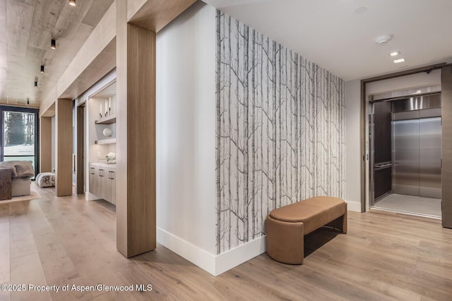 corridor featuring elevator, baseboards, an accent wall, and wood finished floors
