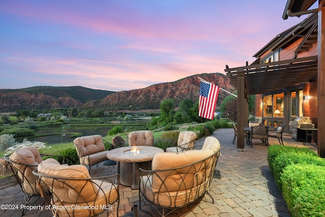 patio terrace at dusk featuring a pergola, a grill, a water and mountain view, and an outdoor living space with a fire pit