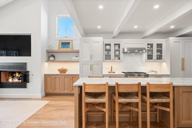 kitchen with a warm lit fireplace, under cabinet range hood, light countertops, beamed ceiling, and a kitchen bar