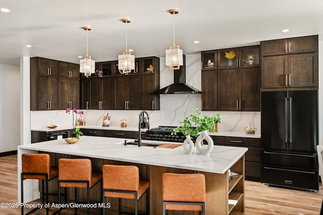 kitchen with decorative light fixtures, an island with sink, sink, high end black fridge, and wall chimney exhaust hood