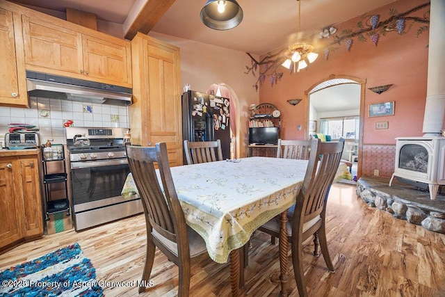 dining space with a wood stove, light wood finished floors, arched walkways, and beam ceiling