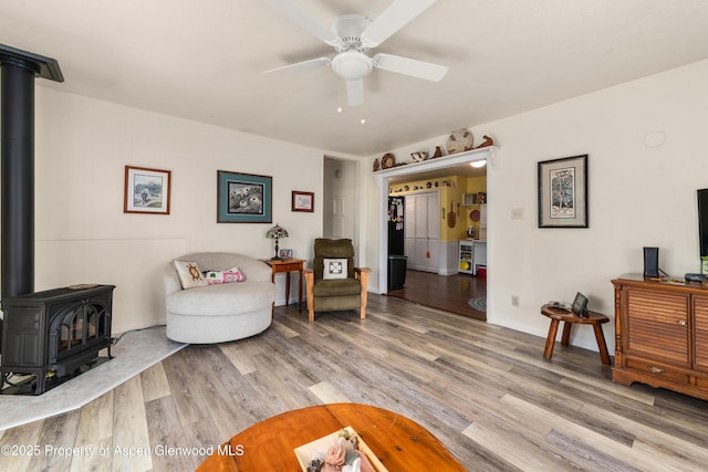 sitting room featuring a wood stove, ceiling fan, light wood-style flooring, and baseboards