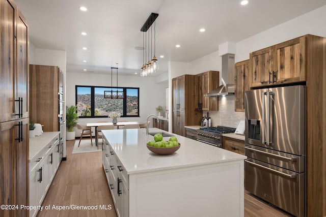 kitchen featuring high end appliances, wall chimney range hood, a center island with sink, white cabinetry, and hanging light fixtures