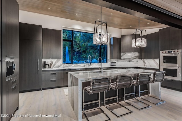 kitchen with wooden ceiling, light stone counters, double oven, decorative light fixtures, and a center island with sink