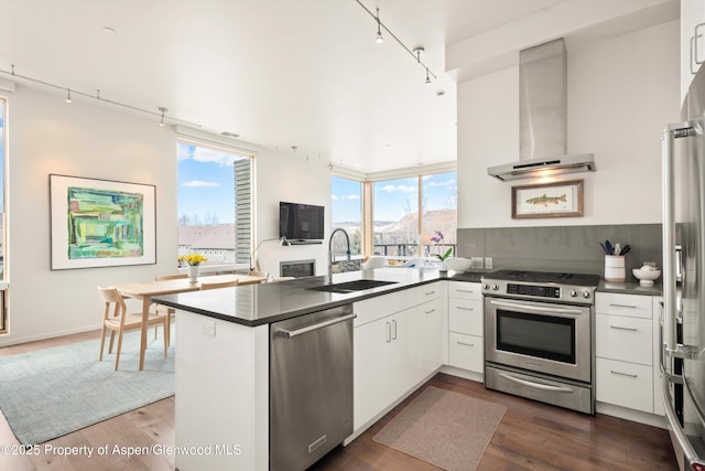 kitchen with dark countertops, appliances with stainless steel finishes, wall chimney range hood, and a sink