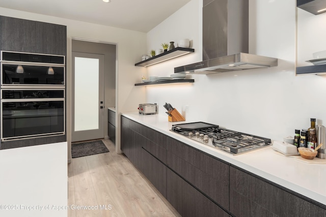 kitchen featuring open shelves, light countertops, stainless steel gas stovetop, wall chimney range hood, and light wood-type flooring