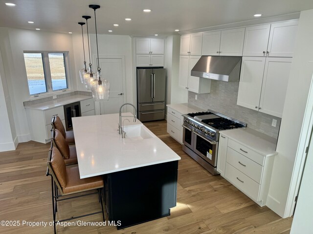 kitchen featuring white cabinets, premium appliances, a kitchen island with sink, and wall chimney exhaust hood