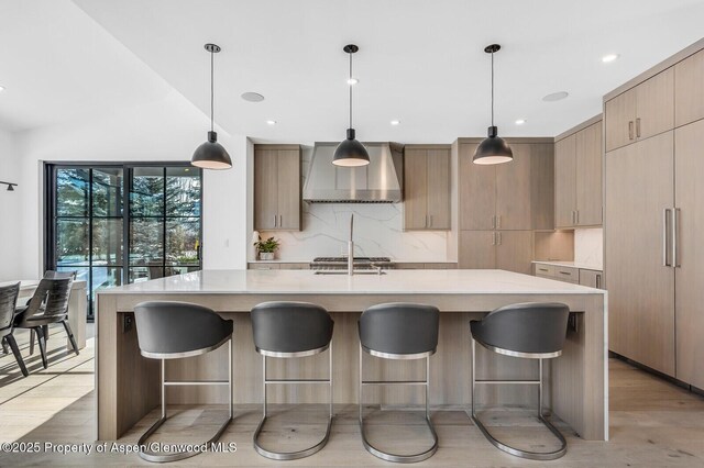 kitchen featuring light brown cabinetry, a center island with sink, wall chimney exhaust hood, and pendant lighting