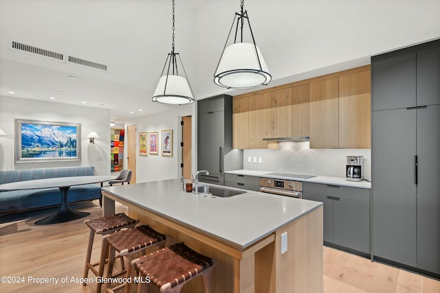 kitchen featuring stainless steel oven, sink, an island with sink, decorative light fixtures, and gray cabinets