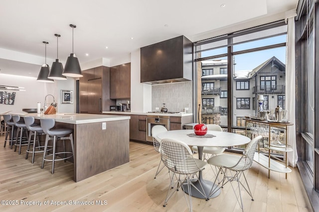 kitchen with hanging light fixtures, light hardwood / wood-style floors, an island with sink, and wall chimney exhaust hood