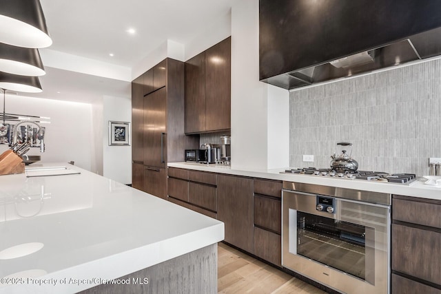 kitchen with light hardwood / wood-style flooring, backsplash, dark brown cabinets, stainless steel appliances, and wall chimney exhaust hood