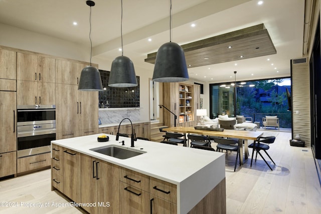 kitchen featuring a center island with sink, sink, light wood-type flooring, decorative light fixtures, and stainless steel double oven