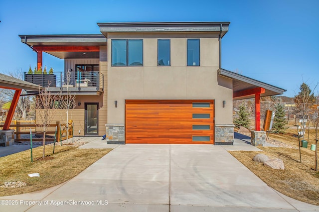 contemporary house featuring an attached garage, a balcony, driveway, stone siding, and stucco siding