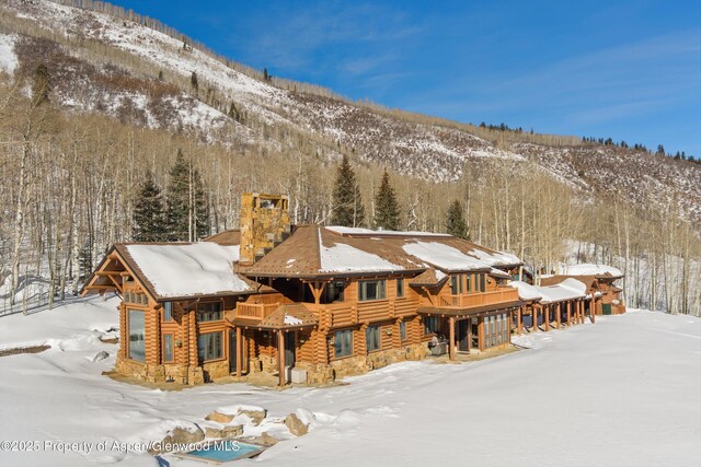 snow covered house featuring a chimney, a forest view, a mountain view, and log siding