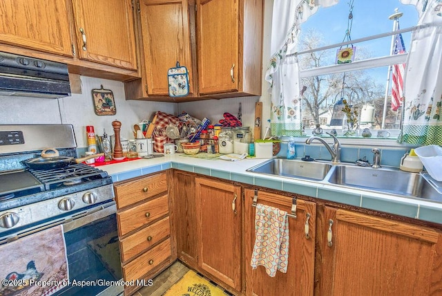 kitchen with tile counters, under cabinet range hood, brown cabinets, stainless steel gas range, and a sink