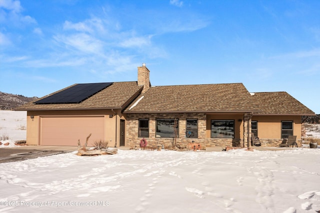 single story home with a garage, stone siding, roof mounted solar panels, a chimney, and stucco siding