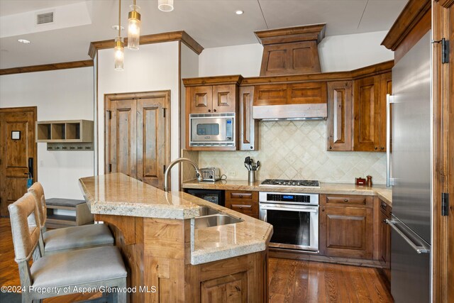 kitchen with sink, hanging light fixtures, tasteful backsplash, built in appliances, and a kitchen island with sink