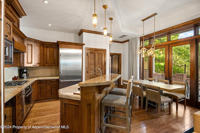 kitchen with tasteful backsplash, light stone counters, built in appliances, decorative light fixtures, and a center island with sink