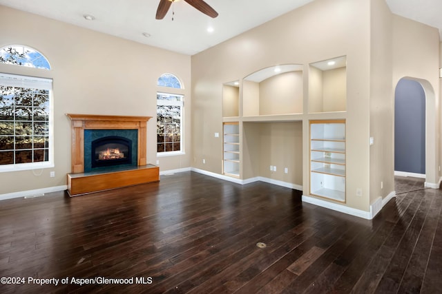 unfurnished living room featuring built in shelves, ceiling fan, dark wood-type flooring, a high ceiling, and a tiled fireplace