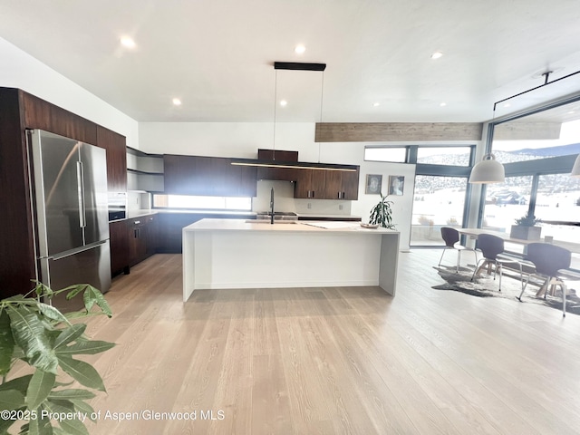 kitchen with pendant lighting, stainless steel fridge, dark brown cabinetry, light hardwood / wood-style floors, and a center island with sink