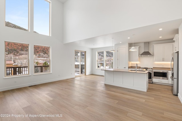 kitchen featuring white cabinetry, wall chimney exhaust hood, hanging light fixtures, stainless steel appliances, and a center island with sink