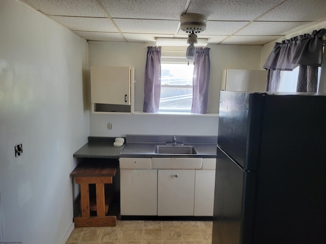 kitchen with black fridge, a drop ceiling, white cabinetry, light tile floors, and sink