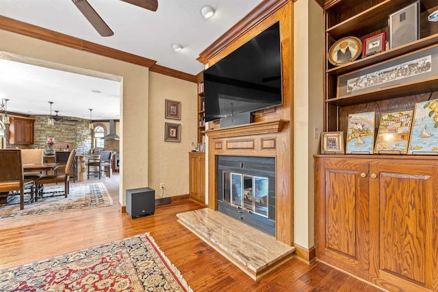 living room with ceiling fan, crown molding, light wood-type flooring, and a stone fireplace