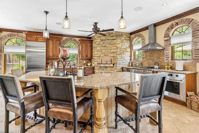 kitchen featuring an island with sink, ceiling fan, stainless steel appliances, wall chimney range hood, and decorative light fixtures