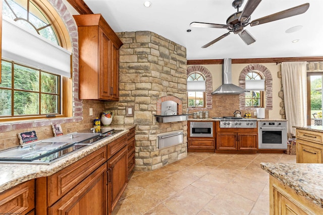 kitchen featuring appliances with stainless steel finishes, tasteful backsplash, light stone counters, ceiling fan, and wall chimney exhaust hood