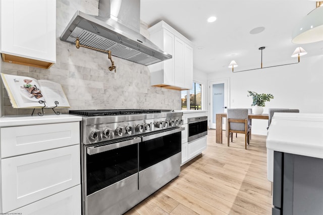 kitchen with appliances with stainless steel finishes, white cabinetry, backsplash, hanging light fixtures, and wall chimney range hood