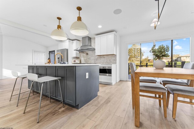 kitchen featuring pendant lighting, oven, white cabinets, wall chimney range hood, and a center island with sink