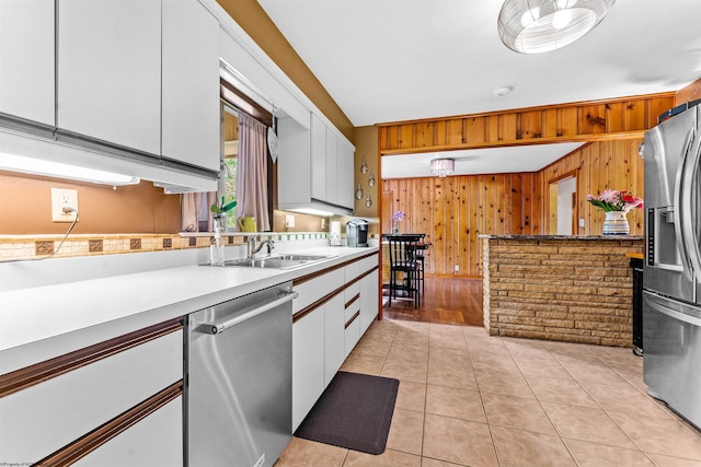 kitchen featuring sink, wooden walls, appliances with stainless steel finishes, light tile patterned flooring, and white cabinetry